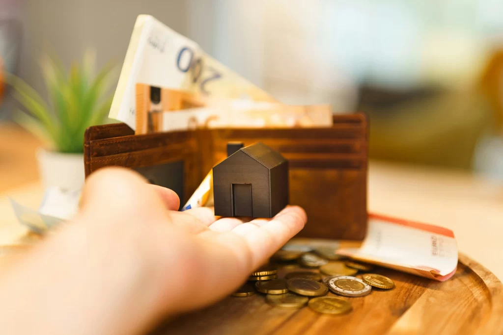 Una mano sostiene un casa pequeña negra, de fondo, una billetera con dinero desordenado.