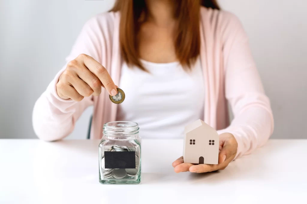 Mujer sostiene una casa de madera mientras deja una moneda en un recipiente de vidrio.